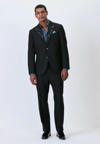 Schwarzer Anzug mit floralem Stickmuster am Revers, kombiniert mit einem blauen Hemd. Glänzende schwarze Schuhe runden den formellen Look ab.