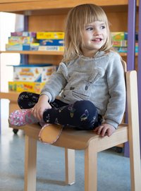 Kind sitzt auf einem Holzstuhl, trägt einen grauen, flauschigen Kapuzenpullover mit Hasenmerkmalen und gemusterte Leggings, dazu rosafarbene Schuhe. Klassenzimmer-Hintergrund.