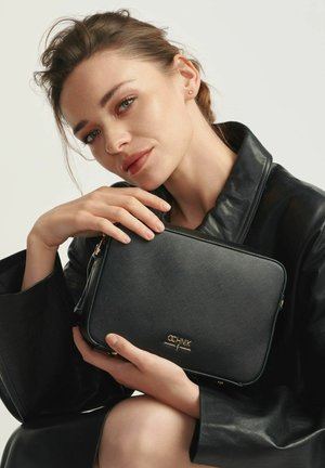 Czarna torebka z fakturowanej skóry z złotymi akcentami, z zamknięciem na zamek błyskawiczny, trzymana przez osobę w czarnym skórzanym płaszczu, na neutralnym tle.