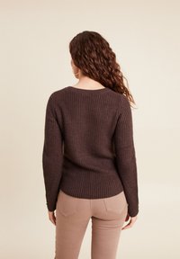 Brauner Strickpullover mit geripptem Muster und langen Ärmeln, kombiniert mit hellbraunen Hosen. Sichtbare Nahtdetails an Ärmeln und Taille.