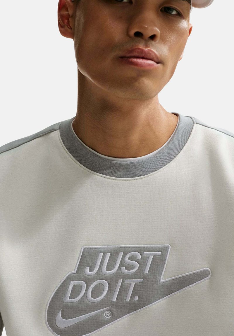 Joven con sudadera blanca y gris con un gran logo en relieve "JUST DO IT." de Nike en el pecho y una gorra blanca.