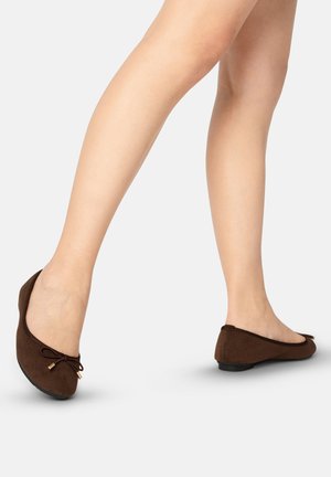Jambes portant des ballerines marron avec de petits nœuds décoratifs, montrées en train de marcher sur un fond clair uni.