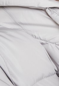 Světle šedá pufovaná bunda má hladký povrch s jemným švem a záhyby, což naznačuje izolaci a teplo.