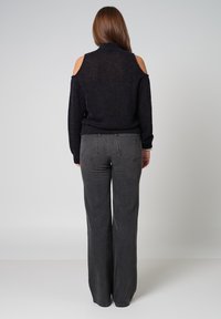 Schwarzer Cold-Shoulder-Pullover mit hohem Kragen, kombiniert mit dunkelgrauen, hochgeschnittenen, weit ausgestellten Jeans, die Rückentaschen und dezente Nähte aufweisen.