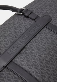 Sac gris foncé avec un motif texturé, doté d'une anse en cuir noir embossée avec "MICHAEL KORS". Accents de quincaillerie lisses visibles.