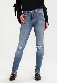 Jeans skinny de denim azul con un acabado desteñido, que presentan desgarros en las rodillas y un diseño de cintura alta. Llevados con mocasines negros.