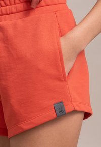 Pantaloni corti in cotone arancioni con vestibilità rilassata, dotati di una tasca laterale e una toppa in tessuto grigia con icona delle forbici sull'orlo.