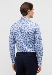 Witte shirt met een blauw bloemmotief, lange mouwen en een button-down kraag. Gedragen met donkere broeken, toont een getailleerde pasvorm.
