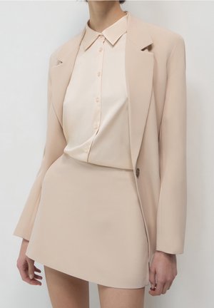Vrouw die een beige blazer draagt, bijpassende minirok en een lichtbeige satijnen blouse met knopen tegen een effen witte achtergrond.