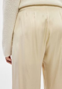 Άτομο που φοράει κρεμ σατέν παντελόνι και μπεζ πλεκτό πουλόβερ, εμφανιζόμενο από τη μέση μέχρι τη μέση μηρών, στέκεται με το ένα χέρι �χαλαρό.