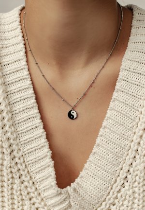 Silberne Kettenhalskette mit einem runden schwarz-weißen Yin-Yang-Anhänger. Trägt man über einem strukturierten weißen Strickpullover mit V-Ausschnitt.