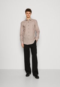 Camicia a maniche lunghe di colore marrone e bianco con motivo, con chiusura a bottoni, abbinata a pantaloni neri a gamba larga e scarpe nere.