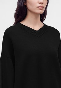 Femme aux longs cheveux foncés portant un pull noir à col en V sur un fond clair uni, montrant le haut du torse et le bas du visage.