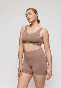 Mujer con cabello rubio recogido en una coleta, vistiendo un conjunto deportivo beige que combina, compuesto por un sujetador deportivo y pantalones cortos de talle alto, sobre un fondo gris liso.
