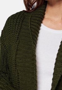 Stickad kofta i mörkgrön, med en strukturerad, ribbad design och öppen framsida, lager på lager över en vit topp med rund halsringning.