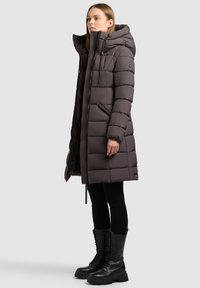 Podstavljeni kaput do koljena u tamnosivoj boji, s kapuljačom, patentnim zatvaračem sprijeda i bočnim džepovima. Uparen s crnim čizmama na vezivanje i tajicama.