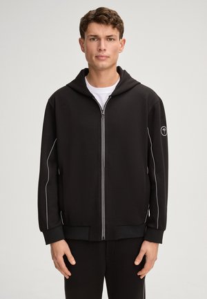 Sweatshirt à capuche noir en tissu doux, avec une fermeture éclair sur le devant, des accents blancs sur les côtés et un logo sur la manche gauche.
