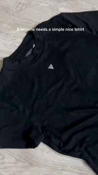 Μαύρο T-shirt με κοντά μανίκια, απλωμένο σε επίπεδη επιφάνεια από ανοιχτό ξύλο, με μικρό λευκό τρίγωνο λογότυπο στο στήθος και κείμενο πάνω από αυτό.