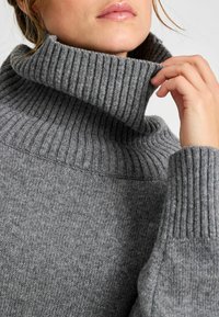 Maglione grigio lavorato a maglia con un alto collo a coste e maniche lunghe. La texture è morbida e liscia, con sottili variazioni di tonalità.