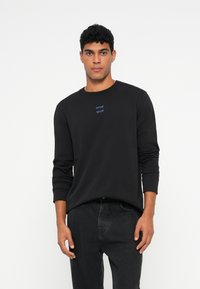 Vīrietis ar melnu garām piedurknēm kreklu, uz kura kr�ūtīm ir neliels zils teksts, un tumšām džinsām, stāv uz vienkārša balta fona.