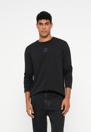 Mann in einem schwarzen Langarmshirt mit kleinem blauen Text auf der Brust und dunklen Jeans, der vor einem schlichten weißen Hintergrund steht.