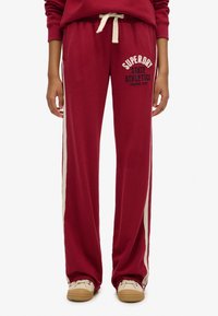 Bordeaux sweatpants med en cremegul stripe ned langs siderne. Har et broderet logo og en snøre i taljen. Fremstillet i blødt, tekstureret stof.