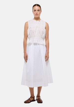 Femme debout face à l’avant portant un chemisier blanc sans manches, une jupe midi blanche et des sandales plates tressées marron sur un fond uni.