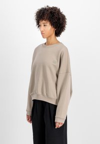 Sudadera beige de corte relajado, con puños y dobladillo acanalados, que presenta un pequeño parche con logo en el pecho. Tejido suave y texturizado.
