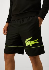 Calções pretos com cintura elástica e bolsos laterais. Apresentam um logótipo de crocodilo verde e riscas horizontais em verde-neon. Material leve.