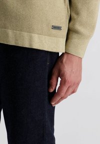 Beige gestrickter Pullover mit einer strukturierten Webart, Rippbündchen und einem kleinen schwarzen Etikett am Saum. Kombiniert mit dunkelblauen Jeans.