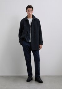 Manteau en laine noir à double boutonnage avec de gros boutons, porté sur une chemise grise et un pantalon sombre. Des chaussures en cuir noir complètent la tenue.