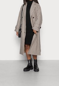 Dubbelknäppt trenchcoat i ett rutigt mönster i beige och brunt, kombinerat med en svart polokragsklänning och kraftiga svarta ankelkängor.