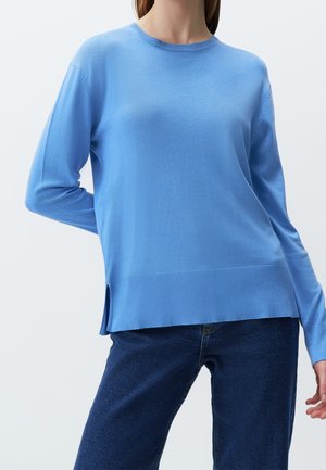 Femme vêtue d'un pull uni bleu clair à manches longues et d'un jean bleu foncé sur un fond uni clair.