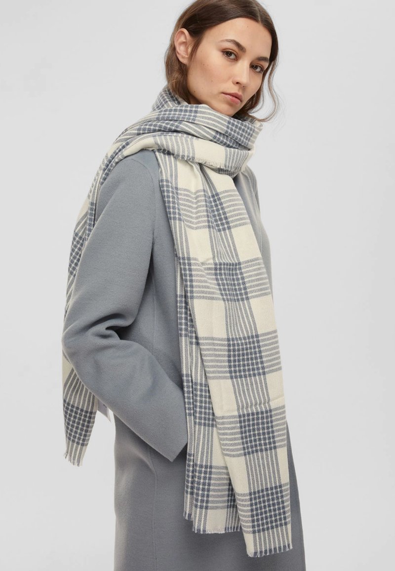 Sciarpa a quadretti leggera in crema e blu navy, realizzata in un tessuto morbido. Presenta un motivo a scacchi e bordi frangiati, appoggiata su un cappotto grigio.