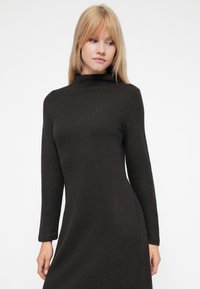 Πλεκτό φόρεμα σε σκούρο γκρι με ψηλό λαιμό, μακριά μανίκια και εφαρμοστή σιλουέτα, διαθέτοντας υφή υφάσματος. Λείοι ραφές και καθαρές γραμμές.