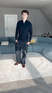 Model wearing a black zip-up sweater and dark, loose-fitting pants stands on a light rug, with brown shoes and a curved, blue sofa in the background.