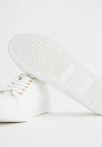 Vita sneakers i canvas med platta snören, guldöglor och en strukturerad gummisula med diamantmönster.