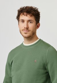 Grüner Sweatshirt mit rundem Halsausschnitt, gerippten Bündchen und einem kleinen braunen Baum-Logo auf der Brust. Weiche Textur und taillierte Passform.