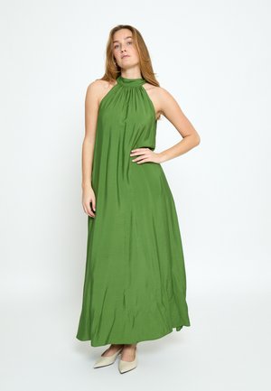 Frau, die in einem ärmellosen, grünen Maxikleid mit Neckholder und weißen, spitz zulaufenden High Heels vor einem schlichten Hintergrund posiert.