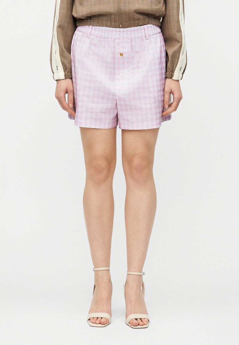 Femme portant un short vichy rose pâle à taille élastique, un haut à manches longues marron et des talons beiges avec bride à la cheville, debout devant un fond blanc.