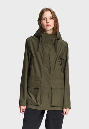 Jeune personne portant une veste à capuche vert olive avec de grandes poches frontales et un pantalon noir, debout devant un fond clair uni.