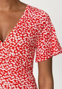 Robe portefeuille à motifs floraux rouges avec des motifs de marguerites blanches. Manches courtes et décolleté en V. Tissu doux et léger avec une texture agréable.