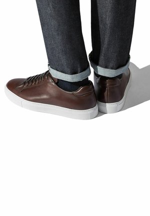 Braune Leder-Sneaker mit niedrigem Profil, weißen Gummisohlen und Schnürdesign, ergänzt durch hochgekrempelte dunkle Denim-Jeans.