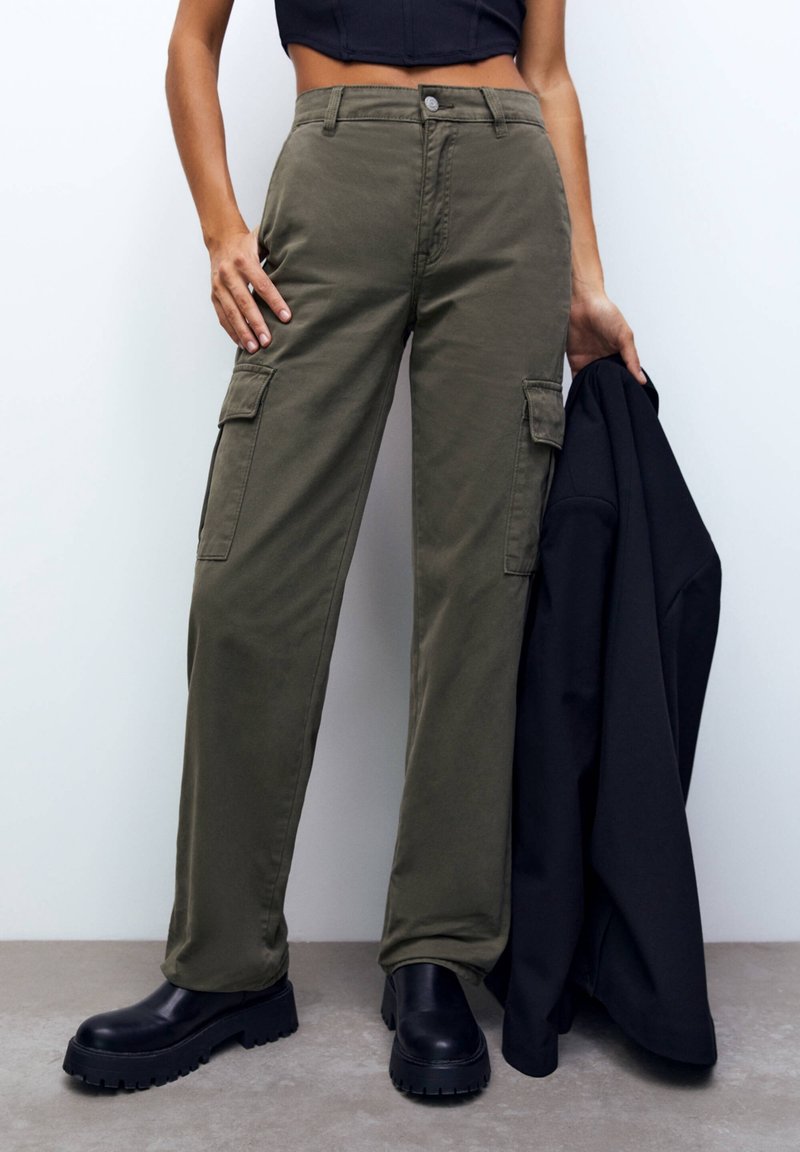Femme portant un pantalon cargo vert olive et des bottes noires épaisses, tenant une veste noire dans une main, debout contre un mur blanc.