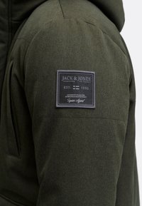 Roheline jakk, millel on tekstuuriga kangas, kapuuts ja õlal ristkülikukujuline brändisilt reljeefse tekstiga.