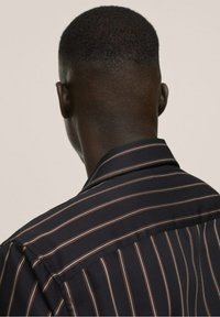 Testa e spalle di una persona con capelli corti e scuri che indossa una camicia nera con strisce verticali marroni e bianche, vista da dietro.