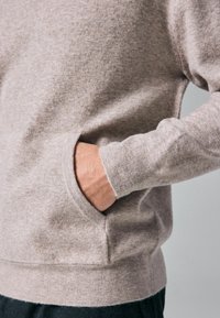 Prawa ręka schowana w beżowej, miękkiej dzianinowej kieszeni, pokazująca ściągacz oraz część czarnych spodni na neutralnym tle.