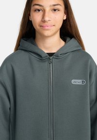 Teenager in einem dunkelgrauen Reißverschluss-Hoodie mit nach unten gezogenem Kapuzen, Logo auf der linken Brustseite, und langen, glatten braunen Haaren vor einem schlichten hellen Hintergrund.