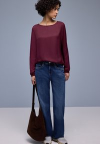 Blouse bordeaux à manches longues associée à un jean bleu à jambes droites et un sac à bandoulière en daim marron. La personne porte des baskets grises.
