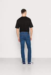 Homem de pé, de frente para uma parede, vestindo uma t-shirt preta, jeans azuis e ténis azul e branco, num fundo simples de parede e chão brancos.